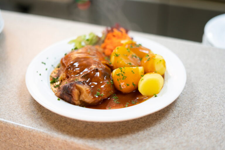 Geschmortes Fleisch mit brauner Sauce, Salzkartöffelchen und Karottensalat auf weißem Teller.