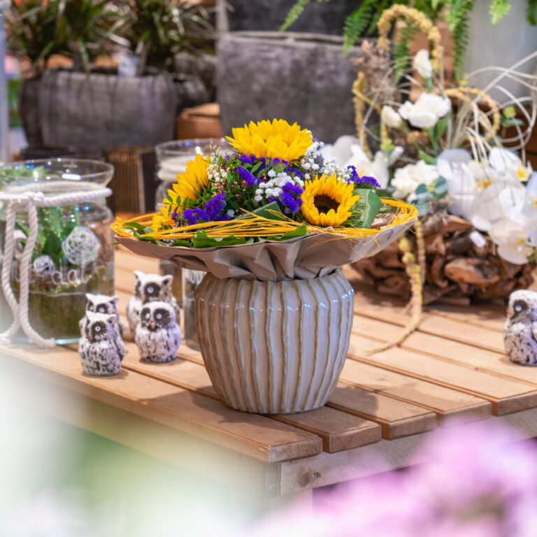 Strauß mit leuchtenden Sonnenblumen und lila Akzenten in einer geriffelten Keramikvase auf Holztisch, Dekoration im Hintergrund.