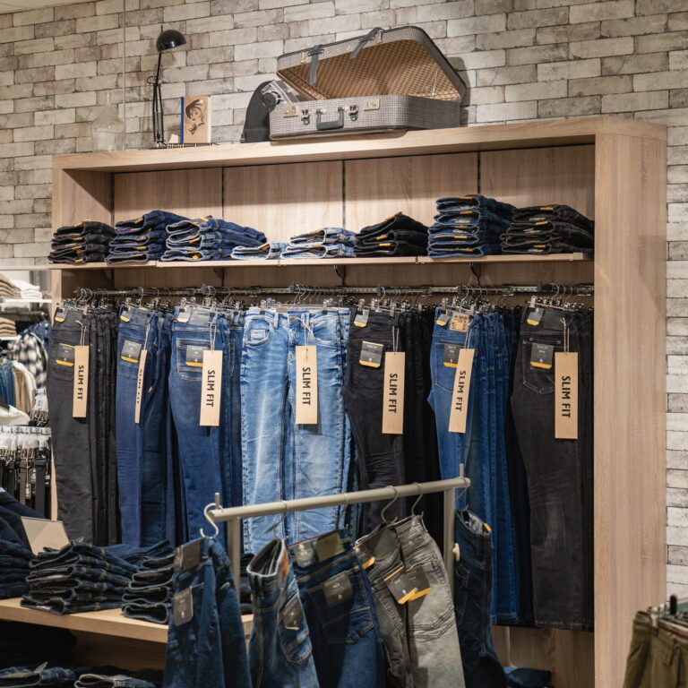Regal mit ordentlich gefalteten Jeansstapeln, aufgehängten Slim-Fit-Jeans und offenem Reisekoffer als Dekoration auf Holzmöbel vor Ziegelwand