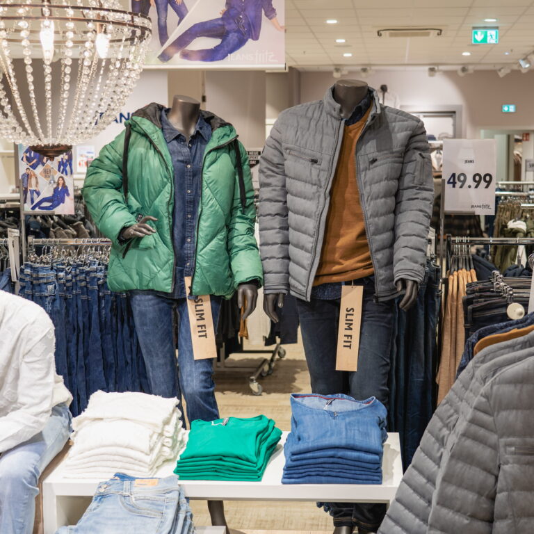 Zwei Kopflose Schaufensterpuppen tragen Winterjacken vor Gestellen mit Jeans und Preisschild 49,99 im Bekleidungsgeschäft.