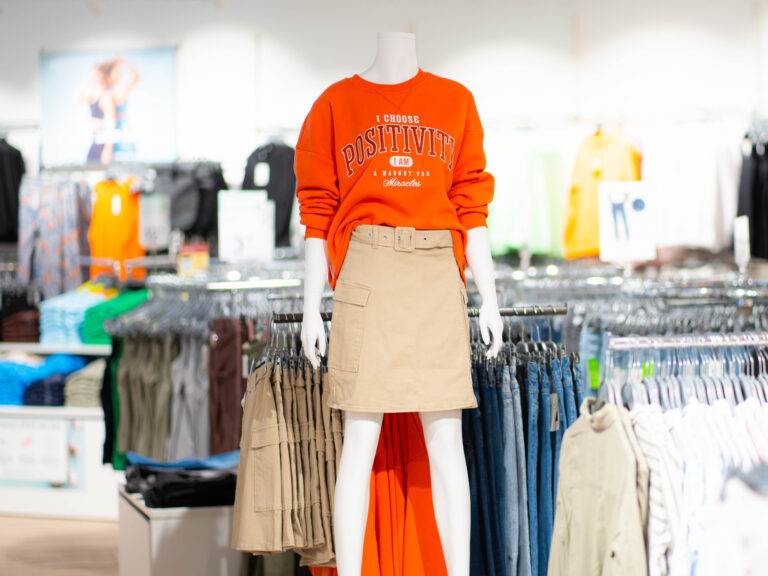 Schaufensterpuppe trägt orangefarbenes Sweatshirt mit Aufschrift „POSITIVITY“ und beiger Gürtelskirt, umgeben von Kleidungsgestellen im Laden.