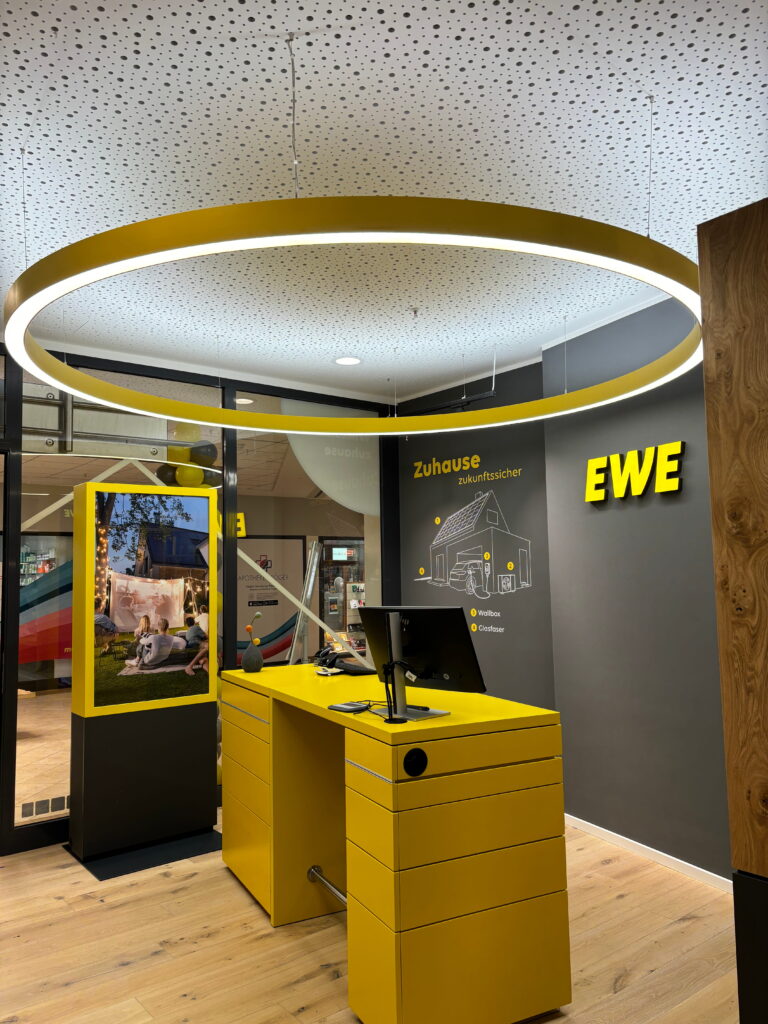 Gelbes Empfangspult mit Monitor unter großem runden Deckenringlicht in moderner Verkaufsfläche mit EWE-Logo an der Wand.