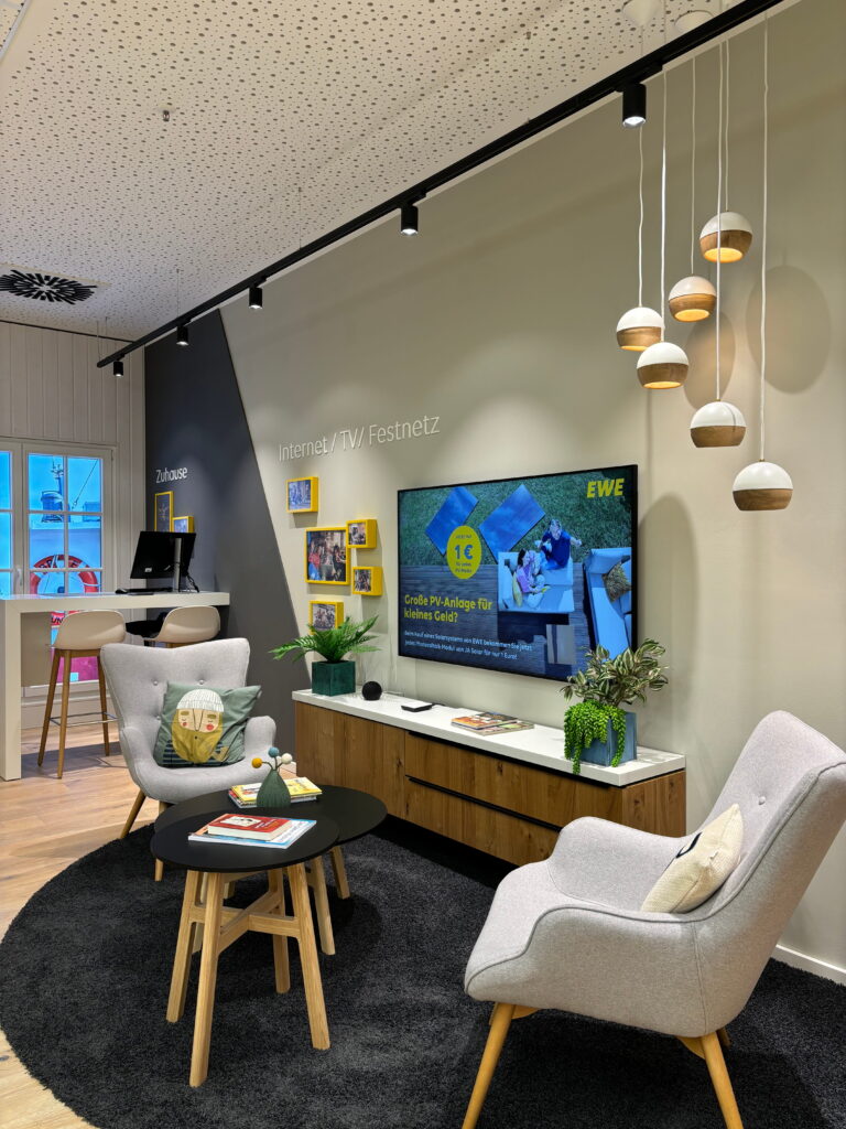 Gemütliche Ladenlounge mit zwei grauen Sessel, runden Kaffeetischen auf schwarzem Teppich, Holzsideboard, TV an der Wand und hängenden Kugelpendeln.