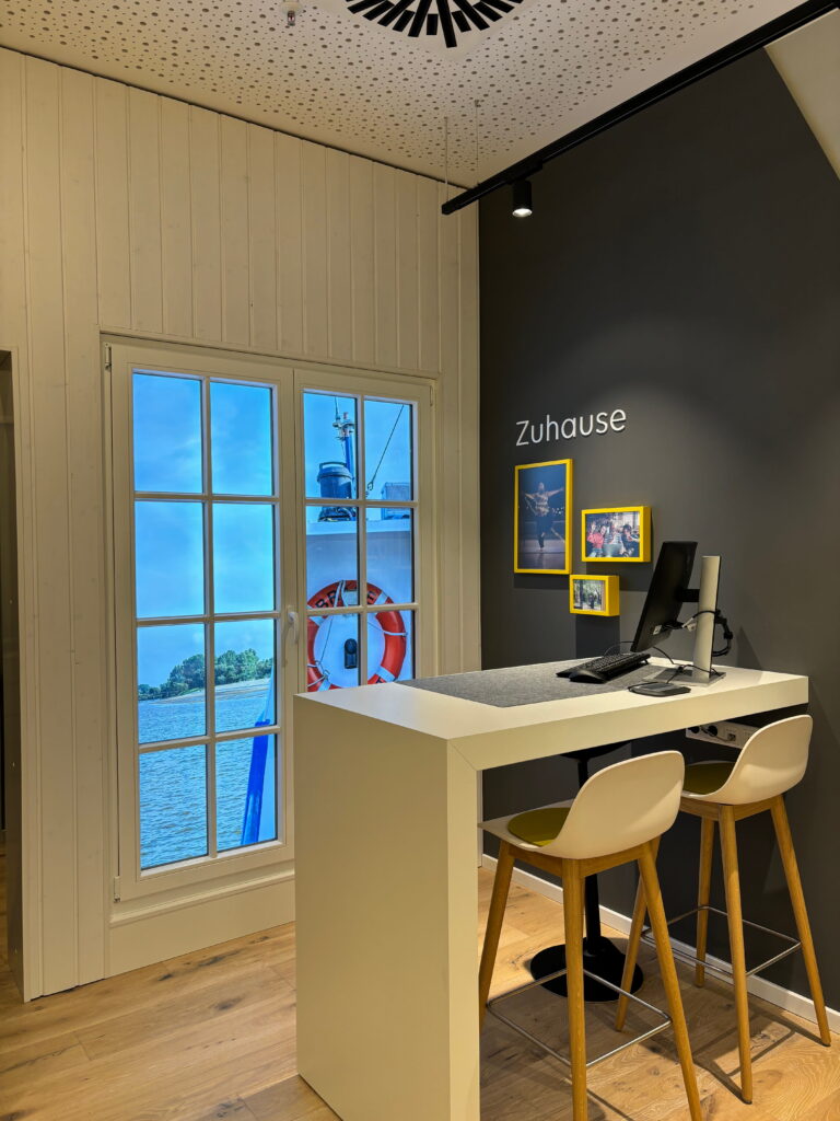 Heller Empfangsbereich mit Stehtisch, zwei Barhockern, Computer, dunkelgrauer Wand mit Schriftzug "Zuhause" und Fenster mit Meeresblick.