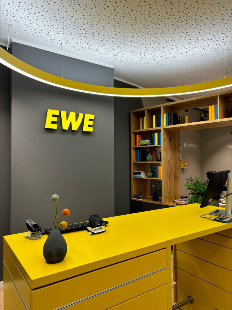 Gelbe Empfangstheke und hängende Leuchtschelle vor grauer Wand mit gelbem EWE-Logo, rechts Holzregal mit bunten Büchern und Schreibtischbereich.