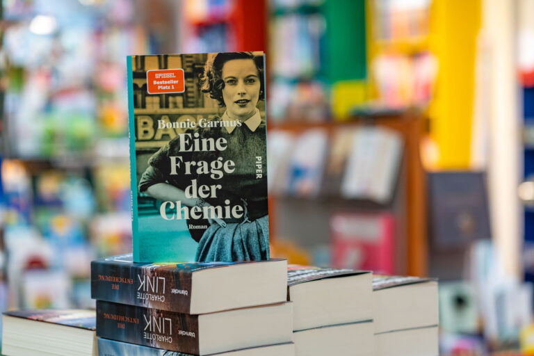 Buchcover von "Eine Frage der Chemie" von Bonnie Garmus mit Schwarzweiß-Porträt einer Frau, im Vordergrund aufgestapelte Bücher in einer Buchhandlung