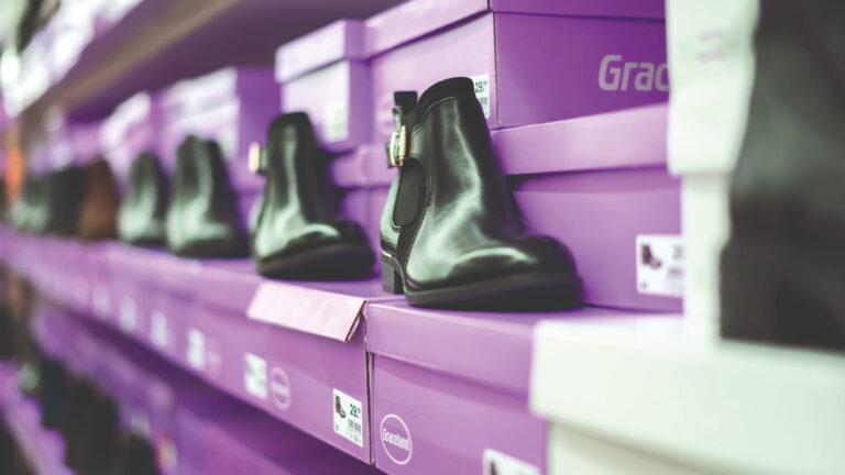 Schwarz glänzende Chelsea-Boots auf gestapelten violetten Schuhkartons in einem Ladenregal, Reihen unscharf im Hintergrund.