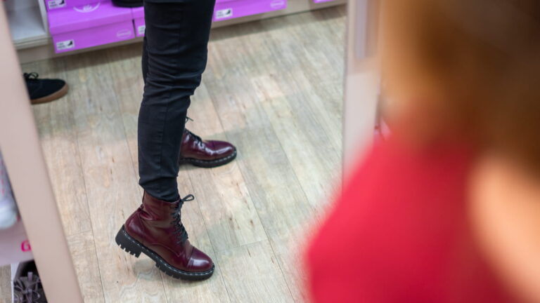 Person trägt schwarze enge Jeans und dunkelrote, glänzende Schnürstiefel mit grober Profilsohle auf Holzfußboden in Ladenumgebung.