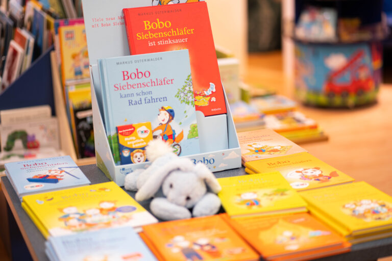 Kinderbücher von "Bobo Siebenschläfer" in buntem Display, im Vordergrund ein Plüschtier und mehrere gelbe und blaue Ausgaben sichtbar