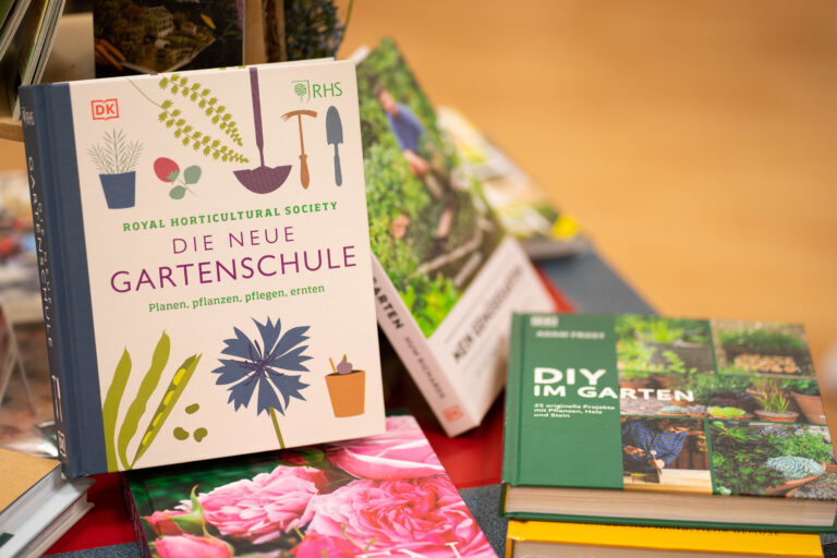 Bücherstapel mit Gartenratgebern, vorn ein Exemplar mit Titel "Die neue Gartenschule" und bunten Pflanzen- und Werkzeugillustrationen.