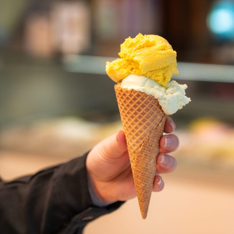 Waffelhörnchen mit zwei Kugeln Eis, oben leuchtend gelb, unten hellcremig, in einer Hand gehalten vor unscharfem Hintergrund