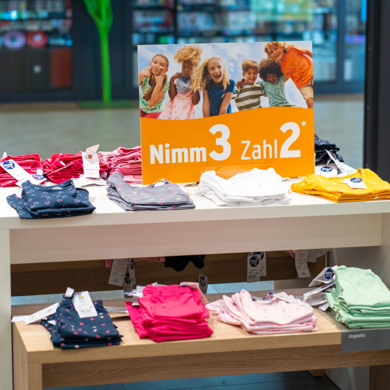 Kinderkleidung auf einem Verkaufstisch mit orangefarbenem Werbeschild "Nimm 3 Zahl 2" und gestapelten bunten Shirts und Hosen.