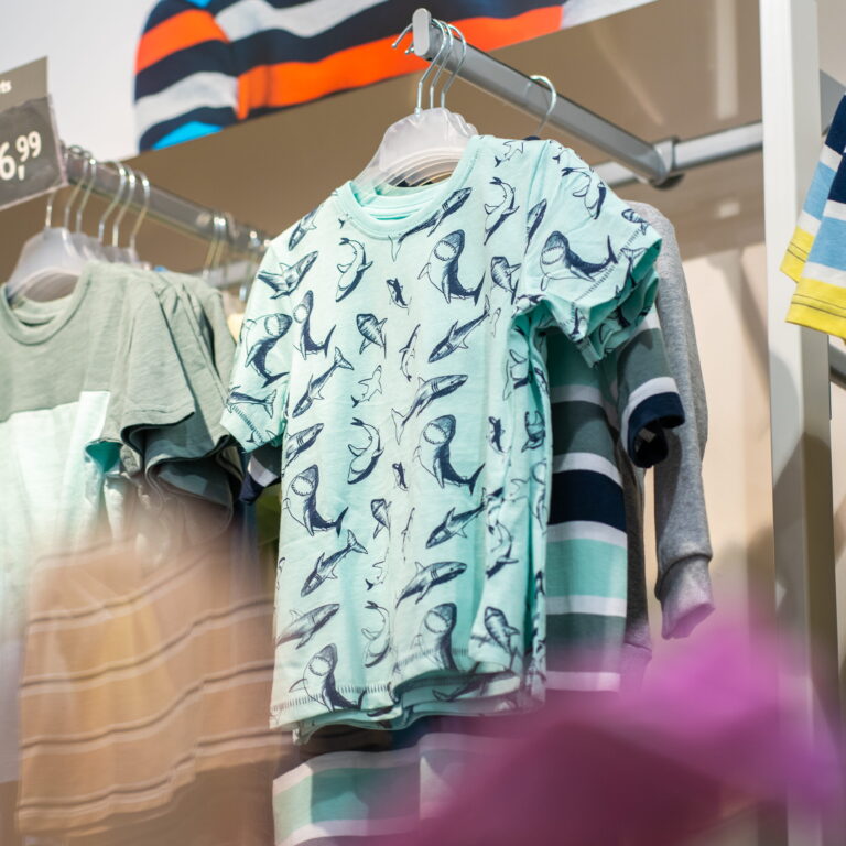 Hellblaues Kinder-T-Shirt mit dunklen Haifischen als Muster, aufgehängt an Kunststoffbügeln in einem Laden.