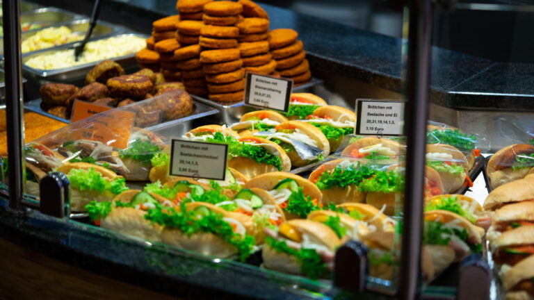 Theke mit belegten Brötchen (Salat, Gurke, Zwiebeln) und gestapelten panierten Frikadellen/Laibchen, Preisschilder sichtbar.
