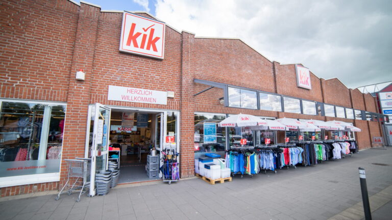 Eingang eines kik-Textilgeschäfts in rotem Backsteingebäude mit offenen Türen, Außenverkaufstischen, Sonnenschirmen und Preisangeboten.