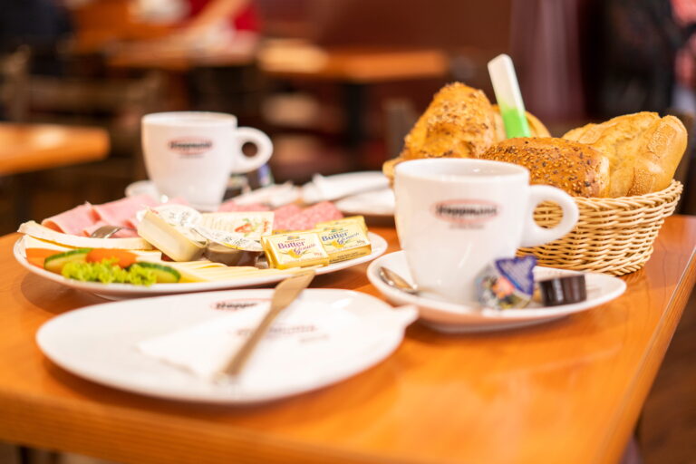 Frühstückstisch mit Kaffee, Teller mit Käse, Wurst und Butterpäckchen sowie Korb mit Brötchen in Café-Atmosphäre