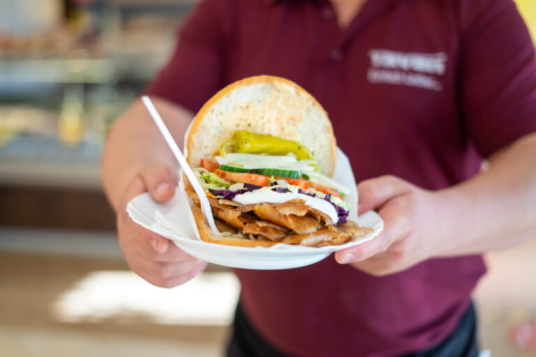 Großes Döner-Sandwich mit Fleisch, Joghurtsoße, Tomaten, Gurken, Salat und Peperoni, präsentiert von einer Person in dunkelrotem Hemd.