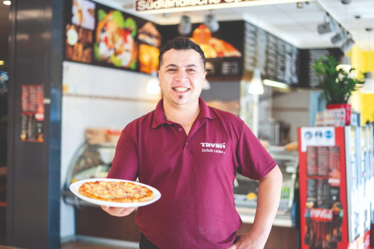 Lächelnder Pizzabäcker in rotem Poloshirt hält eine Pizza auf Teller in einer Imbissküche.