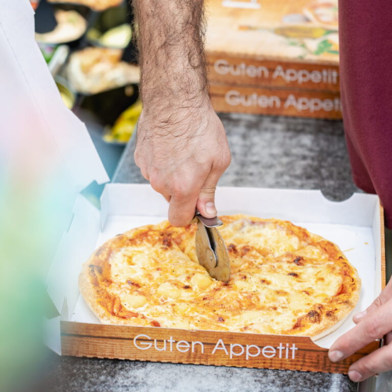 Händiger Arm schneidet mit Pizzaroller eine Käsepizza in einer offenen Pizzaschachtel mit Aufschrift "Guten Appetit".