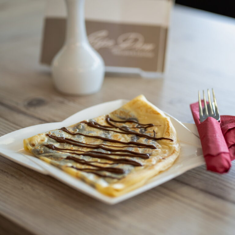 Crepe auf weißem Teller mit Schokoladensauce garniert, daneben rote Serviette und Gabel auf Holztisch