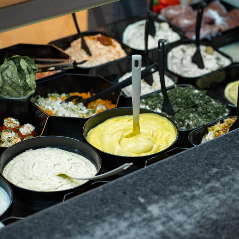 Schalen mit verschiedenen Dips und Salaten; im Vordergrund eine Schale mit hellgelbem Dip und Rührlöffel.
