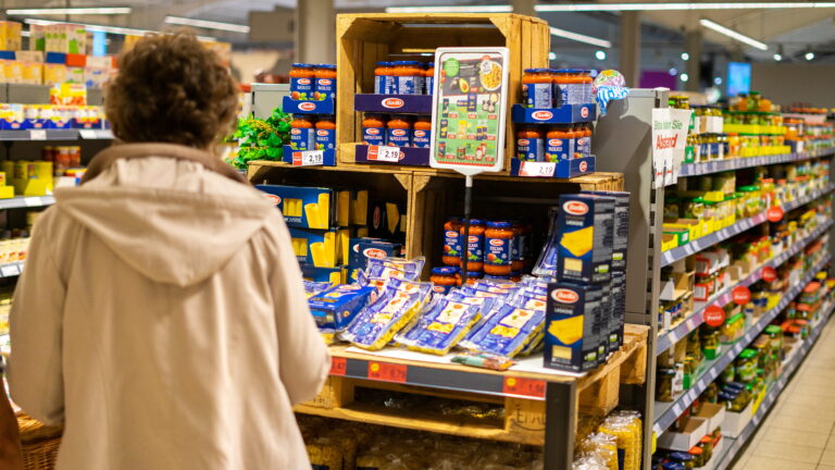 Frau von hinten in hellem Mantel schaut auf Regaldisplay mit Barilla-Pastapäckchen und Tomatensaucen in einem Supermarktgang