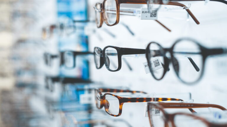 Reihenweise Brillenfassungen verschiedener Formen und Farben auf transparenten Ständern in einem Optikgeschäft, Preisschilder sichtbar.