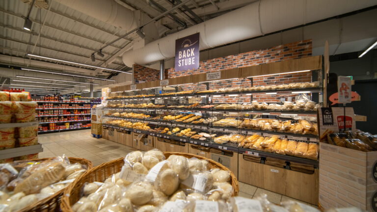 Supermarkt-Bäckerei mit Regalen voller Brötchen und Brot, Körbe mit verpackten Brötchen im Vordergrund und Schild "Backstube" darüber.