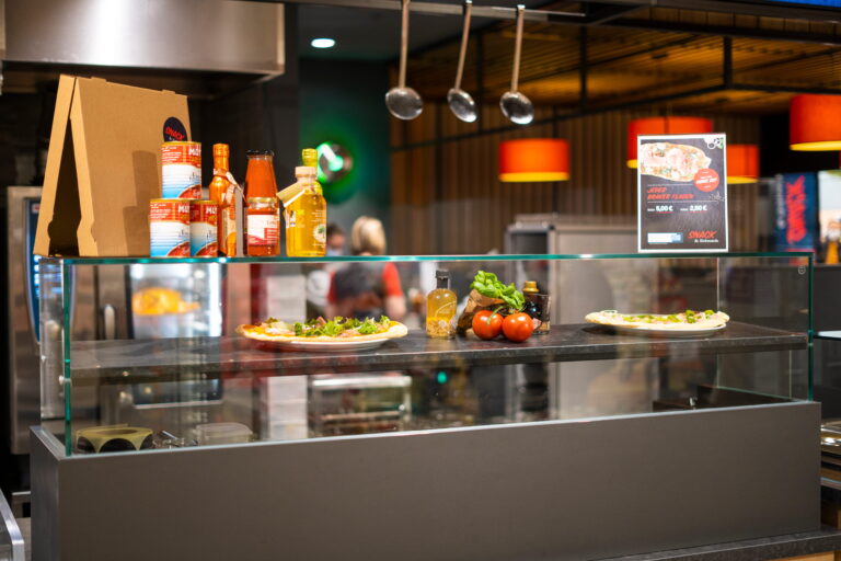 Theke mit zwei belegten Fladenbroten, Tomaten, Ölflaschen, Dosen und Pizzakarton vor Glasvitrine in Imbisslokal