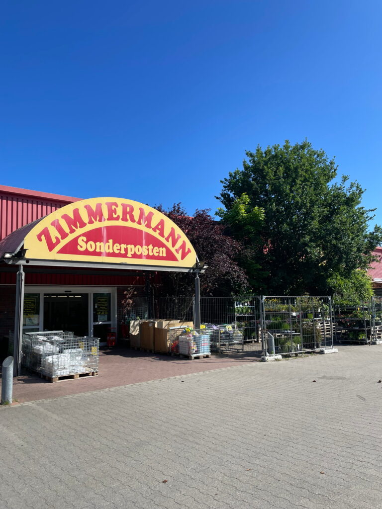 Eingang eines Geschäfts mit rotem Gebäude, gelbem Schild „Zimmermann Sonderposten“, Pflanzen und Metallgitterwaren draußen.