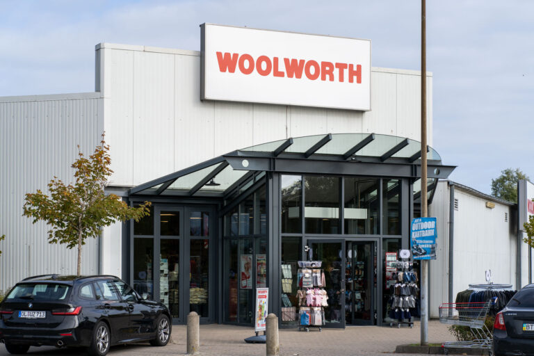Eingang eines Woolworth-Geschäfts mit Glasvordach, Werbeschild oben und Verkaufsständern sowie geparkten Autos davor.